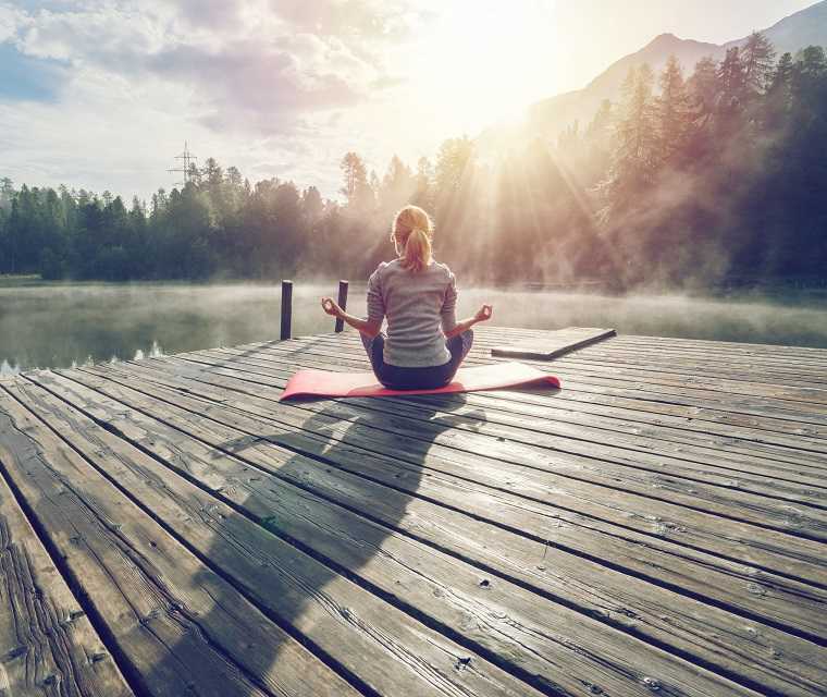 Eine Frau sitzt auf einem Steg und meditiert bei Gegenlicht